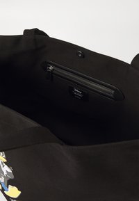 Juodas kanva pirkinių maišas su užtrauktuku viduje, matoma prekės ženklo etiketė ir komiksų stiliaus personažo dizainas išorėje.