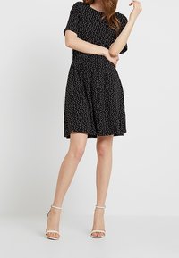 Czarna sukienka z krótkim rękawem w kropki, luźny krój, elastyczna talia i rozkloszowana spódnica, zestawiona z białymi sandałami na obcasie.