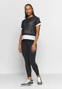 Top desportivo cropped em malha preta sobre uma base branca, combinado com leggings pretas e ténis cinzentos. Apresenta mangas dobradas e detalhes de logótipos.