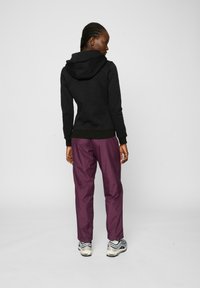 Felpa nera con cappuccio abbinata a pantaloni impermeabili viola. L'outfit presenta polsini a coste e scarpe sportive a contrasto con accenti neri, bianchi e grigi.