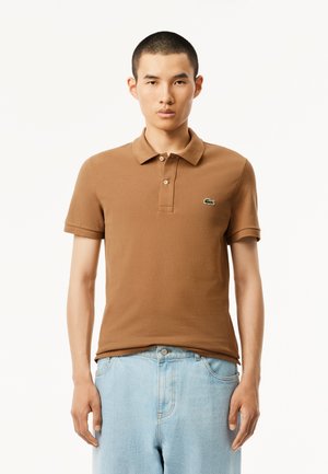 Bruine katoenen polo met korte mouwen, een drieknoops sluiting en een groen geborduurd logo op de borst. Gecombineerd met lichtblauwe jeans.