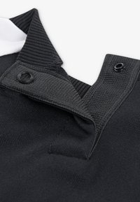 Col polo noir en tissu texturé, avec un col intérieur blanc et deux boutons noirs pour la fermeture.