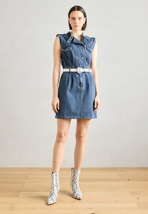 Robe sans manches en jean avec des boutons sur le devant, deux poches poitrine et une ceinture blanche. Portée avec des bottines blanches à imprimé serpent.