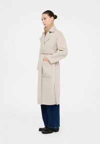 Cappotto in lana beige con colletto a sostegno, tasche laterali e cintura. Indossato sopra un dolcevita a coste e abbinato a jeans a gamba larga e stivaletti.