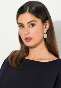 Orecchini color oro con forme rettangolari texture. Il modello indossa una maglietta blu scuro con un motivo a griglia e un ampio scollo. Capelli lisci e dritti.