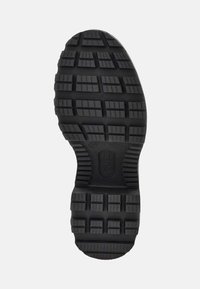Suela de zapato de goma negra con una superficie texturizada y un patrón con surcos profundos y un borde redondeado. Incluye detalle de marca.