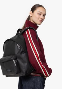 Mochila de tela negra con un bolsillo frontal con cremallera, correas de hombro acolchadas y textura suave, llevada sobre una chaqueta granate con rayas.