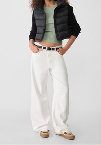 Gilet matelassé noir sur un top vert ajusté, associé à un pantalon large blanc et des baskets beiges. Présente une ceinture noire et des accents de design simples.