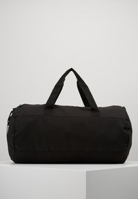 Svart duffelbag med to korte håndtak. Laget av slitesterkt stoff, har den en sylinderform og en glidelås. Enkelt design uten synlige mønstre.