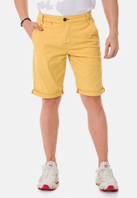 Gelbe Baumwollshorts mit geradem Schnitt, aufgeschlagenen Säumen, kontrastierender roter Stickerei und einem schwarzen Knopfverschluss. Kombiniert mit weißen Sneakern.
