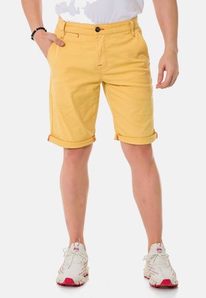 Geel katoenen shorts hebben een rechte snit, opgerolde zomen, contrasterende rode stiksels en een zwarte knoopsluiting. Gepaard met witte sneakers.
