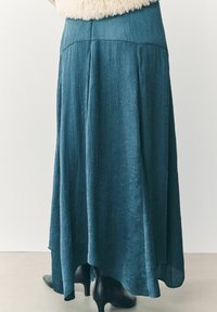 Lunga gonna teal realizzata in tessuto stropicciato, con un design svasato e un orlo asimmetrico, abbinata a stivali neri con tacco.