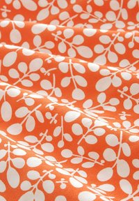 Tissu orange présentant un motif botanique blanc avec des feuilles arrondies et des tiges, créant une apparence texturée et stratifiée.