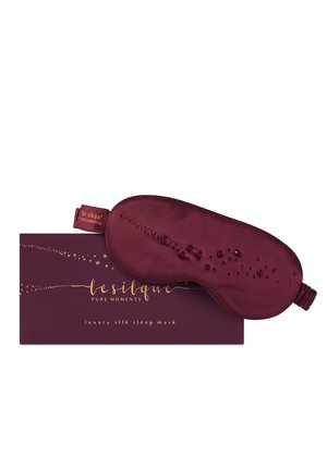 Frau liegt auf einem dunkelvioletten Kissen, trägt eine Satin-Schlafmaske, die über den Augen hochgeschoben ist, und ein helles Satincamisole.