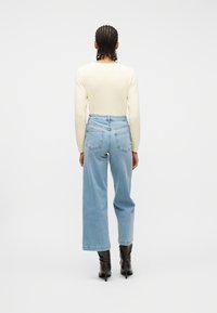 Άτομο με πλεξούδες, φορώντας εκρού πουλόβερ, ανοιχτό μπλε τζιν ψηλόμεσο με φαρδιά μπατζάκια και μαύρες μπότες με τακούνι, στέκεται με την πλάτη προς τα έξω.