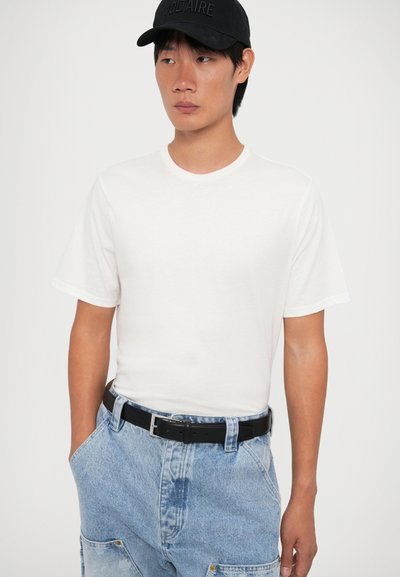 Camiseta de algodón blanca con cuello redondo y mangas cortas, combinada con pantalones de mezclilla azul claro y un cinturón negro. Gorro de béisbol negro con texto en relieve.