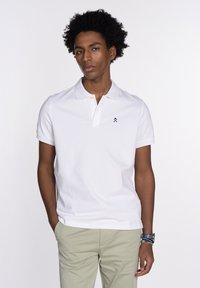 Polo shirt blanco con una tapeta de dos botones, mangas cortas y un logotipo negro en el pecho. La tela parece suave y ligera.