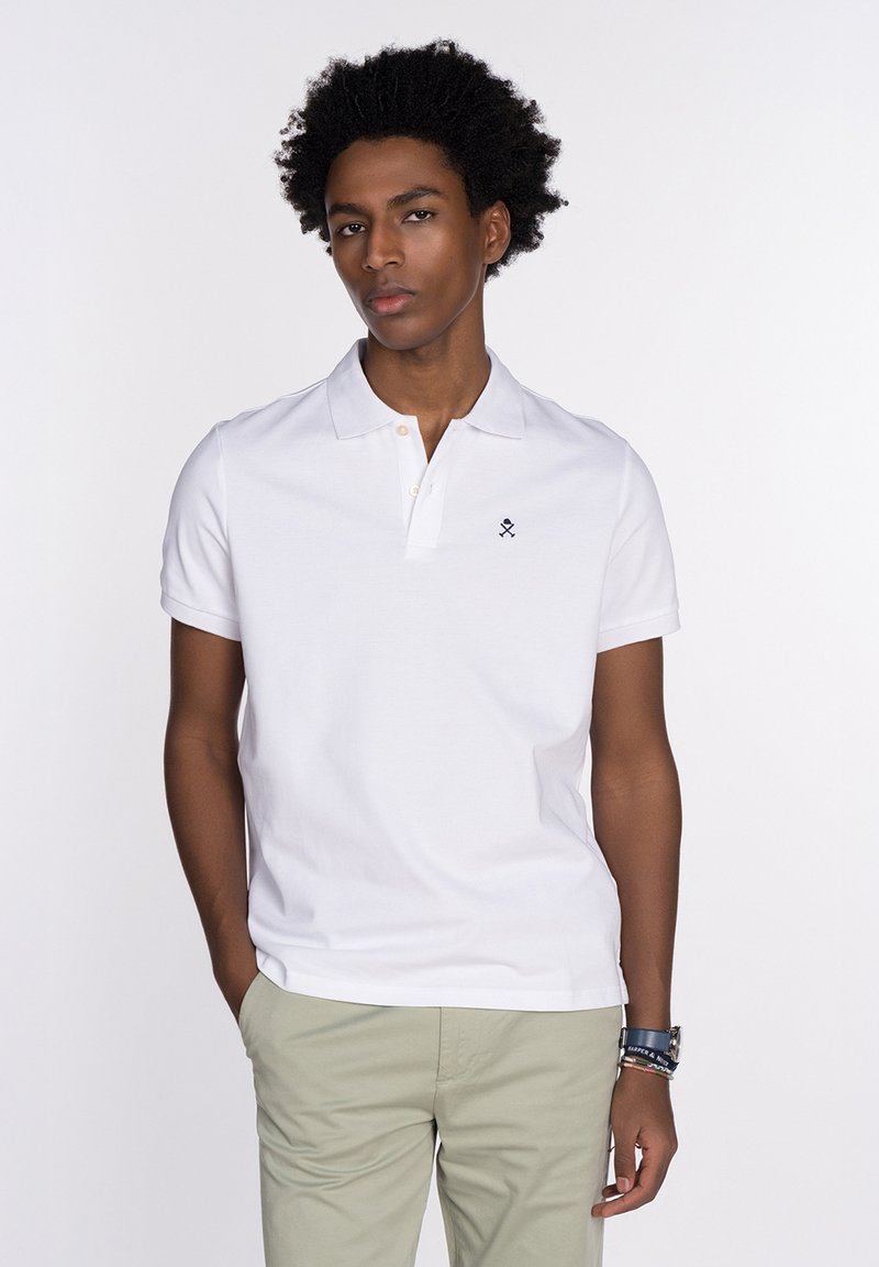 Polo shirt blanco con una tapeta de dos botones, mangas cortas y un logotipo negro en el pecho. La tela parece suave y ligera.