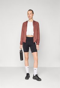 Sportstøj bestående af en cropped hvid top, en maroon letvægtsjakke, sorte cykelshorts med hvide detaljer og sorte sneakers. Holder en vandflaske.