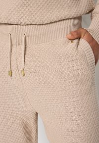 Beige knitted joggers feature a textured pattern, elastic waistband with drawstring, and side pockets with small metal accents.