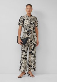 Zwart en beige gemêleerde jumpsuit met korte mouwen, een ceintuur in de taille, wijde benen, gecombineerd met een zwarte clutch met gouden kettingaccenten.