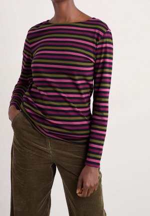 Vrouw draagt een shirt met lange mouwen met paarse, roze en groene strepen en een olijfgroene corduroy broek, poseert met één hand in de zak.