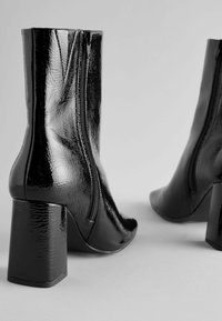 Stivali alla caviglia neri in vernice con chiusura a zip, tacco quadrato e texture liscia. Il design presenta una punta appuntita e una finitura lucida.