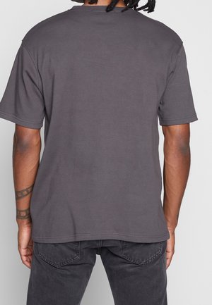 Mand set bagfra iført en ensfarvet mørkegrå kortærmet t-shirt og sorte jeans, med tatoveret underarm synlig.