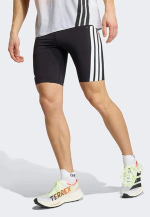 Hombre con pantalones cortos deportivos negros con rayas blancas, calcetines deportivos blancos y zapatillas de running Terrex blancas, de pie con una pierna hacia adelante.