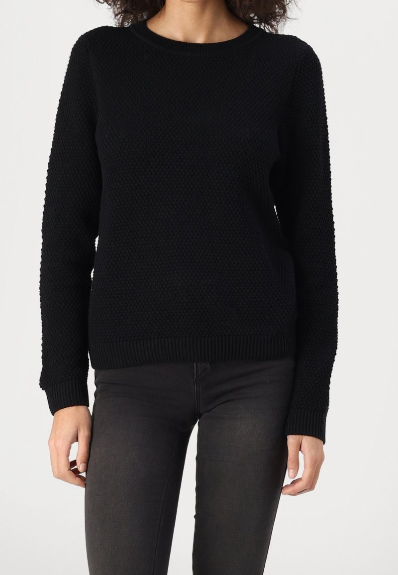 Maglione nero lavorato a maglia con un motivo testurizzato, collo rotondo e polsini a coste. Abbinato a jeans scuri, l'abbigliamento è casual e rilassato.