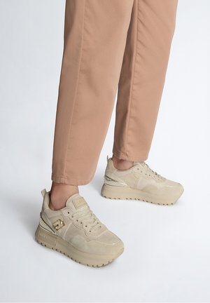 Des pieds portant des baskets compensées beiges et un pantalon marron clair, debout sur une surface blanche unie.