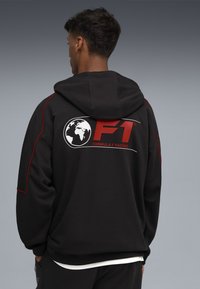 Sort hættetrøje lavet af blødt stof med røde detaljer. På ryggen vises F1-logoet og teksten "FORMULA 1 RACING".