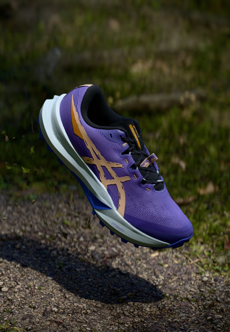 Lilla løbesko med en struktureret overdel, sorte snørebånd og et gyldent logo; hvid mellemsål og blå detaljer på sålen.