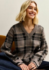 Brown and black checkered v-neck sweater with long sleeves, knit texture, and ribbed cuffs. Worn over dark denim, model is sitting.