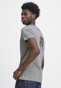 Grå t-shirt med korte ærmer lavet af en bomuldsblanding, prydet med et grafisk design af en person iført briller og hat på ryggen.