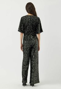 Tuta nera in paillettes con maniche corte, gambe ampie e collo alto. Chiusura con zip sul retro; texture lucida con paillettes su tutta la superficie.