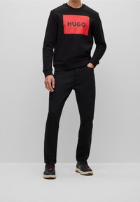 Felpa nera con un logo rettangolare rosso con la scritta "HUGO", abbinata a pantaloni neri e sneakers con dettagli rossi e beige.