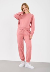Roze sportoutfit van zachte stof, bestaande uit een sweatshirt met een losse pasvorm en taps toelopende joggers met elastische manchetten, gecombineerd met witte sneakers.