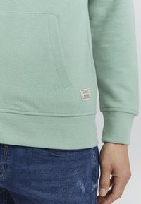 Mintgroene sweatshirt met een buidelzak, geribbelde manchetten en zoom. Beschikt over een vierkant label dat aan de zijkant is genaaid met merkinformatie.