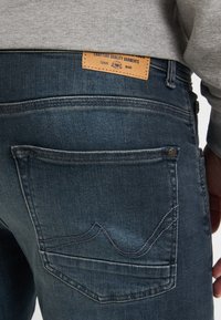 Acercamiento al bolsillo trasero de unos pantalones vaqueros azules con un diseño de olas cosido y una etiqueta de marca de cuero marrón encima de la cintura.