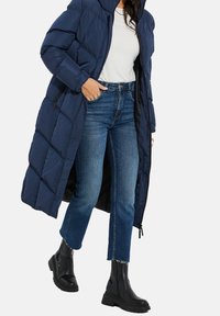 Marineblauwe gewatteerde jas met een quiltontwerp, lange lengte, vooraan met rits en capuchon, gedragen over een wit shirt en gecombineerd met cropped jeans en zwarte laarzen.