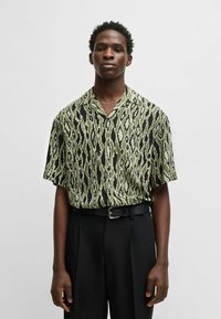 Kurzarmhemd im grün-schwarzen Ikat-Muster, aus leichtem Stoff mit lässiger Passform und einem Eingriffkragen, getragen mit schwarzen Hosen.