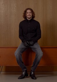 Pull noir zippé, jeans gris, gants noirs et bottines. Homme assis contre un fond en bois, présentant une tenue moderne et simple.