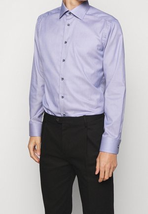 Homme portant une chemise habillée violette claire à boutons, rentrée dans un pantalon formel noir, sur un fond uni.
