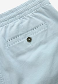 Shorts de culoare albastru deschis, fabricate dintr-un material moale, cu un brâu elastic. Prezintă o buzunar pe spate cu un nasture maro și detalii de cusătură curate.