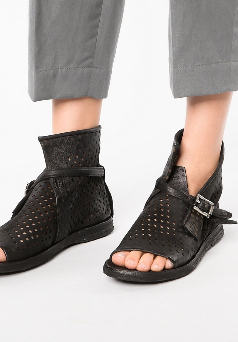 Bottines en cuir noir perforé à bouts ouverts avec des brides réglables à boucle, portées avec un pantalon gris court.