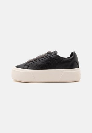 Μαύρο δερμάτινο sneaker χαμηλής κοπής με χοντρή λευκή πλατφόρμα και σκούρα γκρι κορδόνια, πλευρική όψη σε απλό φωτεινό φόντο.