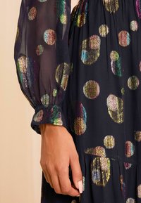 Rochie din țesătură neagră cu buline mari, multicolore, reflective. Mâneci transparente cu mansete strânse. Suprafața texturată adaugă interes vizual.