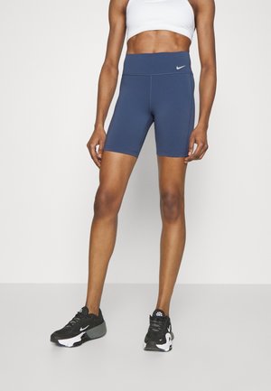 Athletische Person trägt blaue Nike Biker-Shorts mit hoher Taille, weißen Sport-BH und schwarze Nike Laufschuhe und steht auf einer ebenen Fläche.