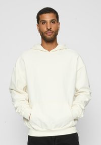 Felpa crema realizzata in tessuto morbido, con tasca frontale, cappuccio con coulisse, vestibilità comoda e polsini a costine. Indossata sopra pantaloni neri.